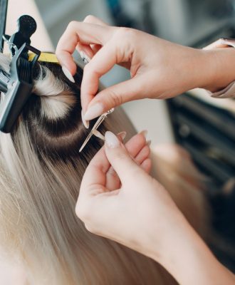 Hairdresser female making hair extensions to young woman with blonde hair in beauty salon.